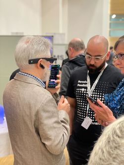 Photo of a group event featuring two men interacting, one wearing smartglasses, holding a smartphone, and another man in a black Envision-branded t-shirt observing closely.