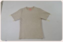 A beige suede T-shirt laid flat on a light gray surface, with the sleeves lightly taped down. The shirt has a soft texture and a pink label inside the collar. The photo looks unedited.