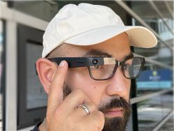 Close-up photo of a man wearing Google Glass smartglasses, touching the side of the device with one finger, wearing a white baseball cap and glasses.